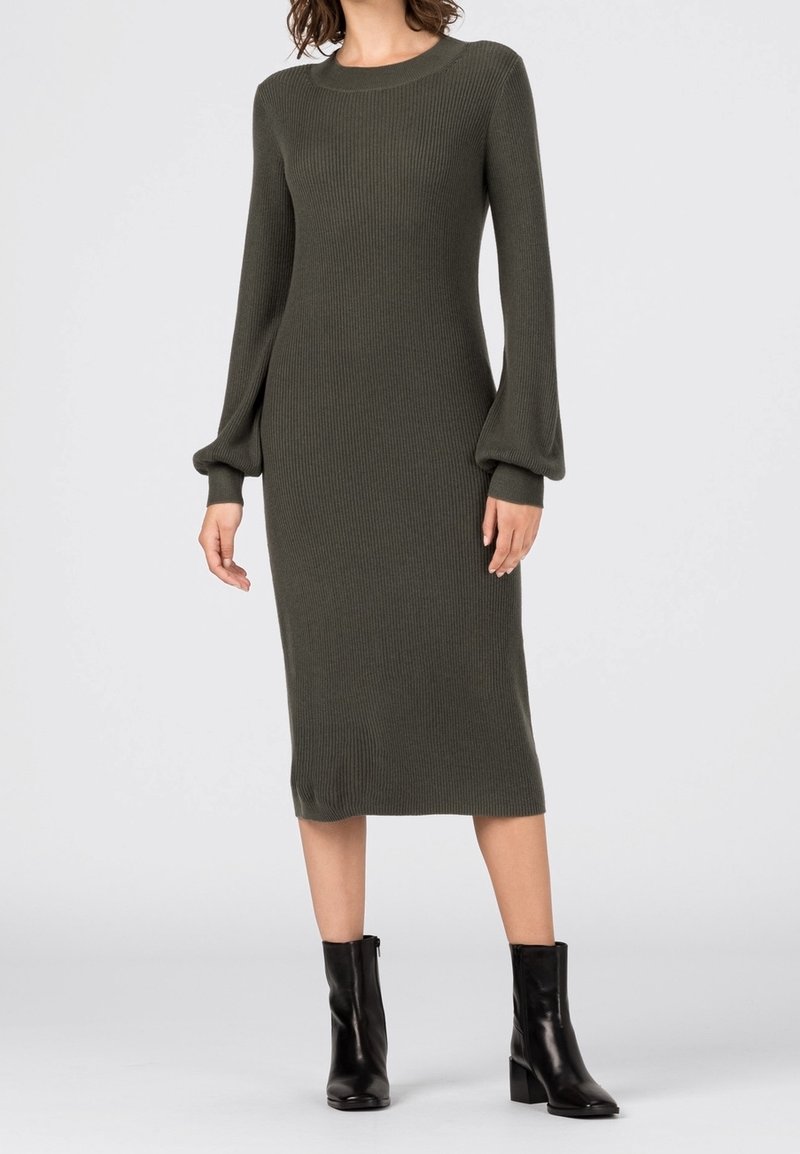 Femme portant une robe pull midi côtelée vert olive avec des manches bouffantes et des bottines noires, debout devant un fond clair uni.
