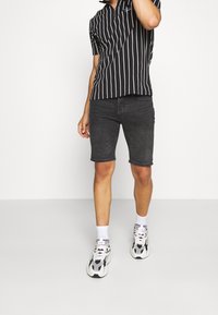 Chemise zippée à manches courtes à rayures noires et blanches, associée à un short en denim foncé. Le modèle porte des baskets blanches et grises avec des chaussettes blanches.