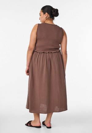Woman with dark hair in bun wearing a sleeveless brown top and matching long skirt, standing barefoot on a white background.