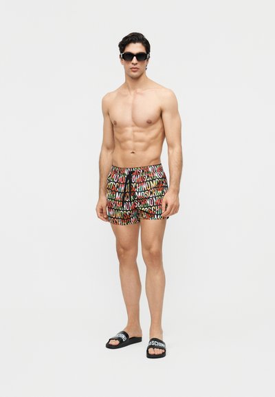 Homme torse nu portant des lunettes de soleil noires, un short coloré avec le logo Moschino, et des sandales à slide noires Moschino, debout devant un fond blanc uni.