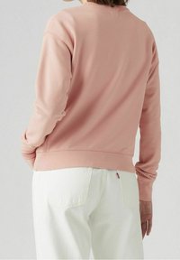 Sudadera rosa de manga larga y escote redondo, combinada con pantalones blancos que tienen bolsillos traseros. Textura suave, diseño sencillo.