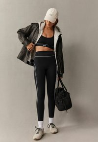 Juodas sportinis crop top'as ir leggingai su kontrastingomis baltomis siūlėmis, derinami su pilku džemperiu ir kreminiu kepurėle, laikant juodą maišelį. Smėliniai sportbačiai.