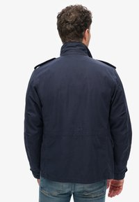 Marineblå, letvægtsjakke med høj krave, skulderepauletter og synlige stingdetaljer, parret med lys blå jeans.