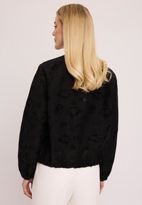 Czarna kurtka o teksturowanej powierzchni z subtelnym wzorem, obszernymi rękawami i marszczonym dołem. Tkanina prezentuje warstwowy design, dodający głębi.