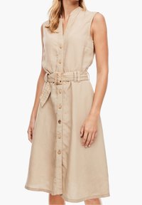 Beige ärmlös klänning i lätt tyg, med en knappslå framtill, bälte i midjan och sidofickor. Subtil texturdetalj.