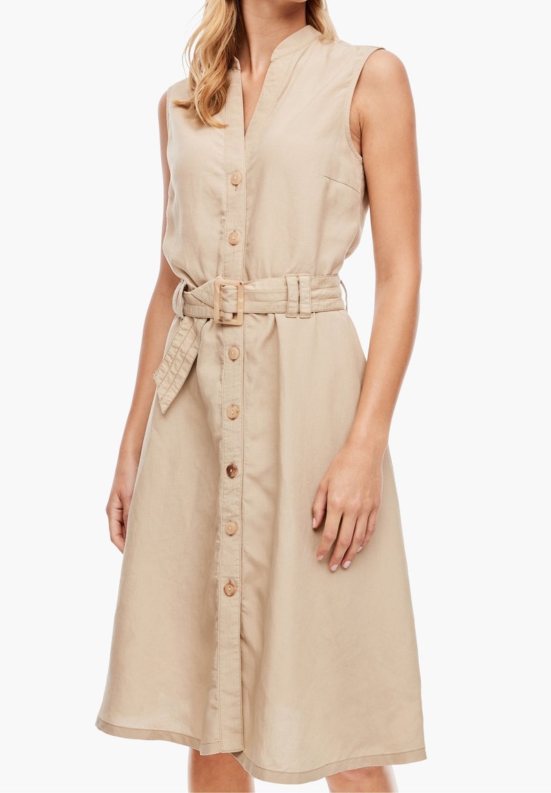 Beige ärmlös klänning i lätt tyg, med en knappslå framtill, bälte i midjan och sidofickor. Subtil texturdetalj.