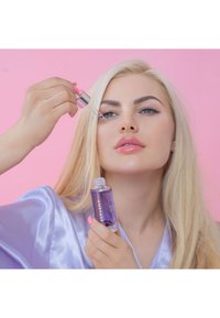 Bottiglia di vetro viola con contagocce, etichettata "AMETISTA." Modella con lunghi capelli biondi tiene la bottiglia, indossando un accappatoio di raso lilla. Sfondo rosa.