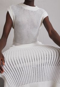 Witte gebreide jurk met opengewerkte textuur, mouwloos ontwerp en geribbelde halslijn. De jurk valt losjes met een vloeiende silhouet.