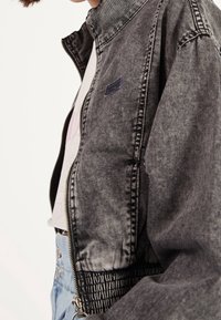 Svart jeansjacka med ståkrage, dragkedjestängning och elastisk midja. Har subtila blekningar och en liten logopatch.