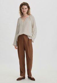 Blusa in lino beige con apertura a chiave e coulisse, abbinata a pantaloni marroni larghi e ballerine marroni. Tessuto liscio, vestibilità rilassata.