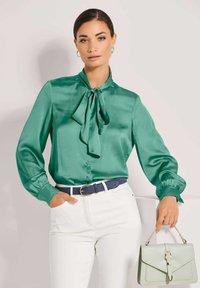 Satinblaue Bluse mit einem Krawattenkragen, langen Ärmeln und Knopfleiste. Kombiniert mit weißen Hosen, einem strukturierten blauen Gürtel und einer hellgrünen Handtasche.