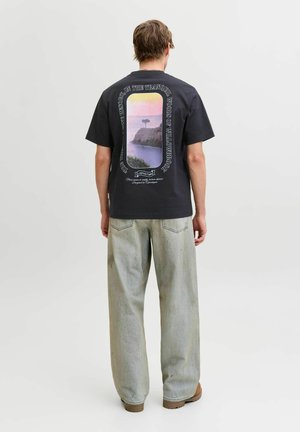 Personne debout vue de dos portant un t-shirt noir avec un motif coloré de falaise et d'arbre, et un jean large délavé.