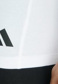 Bílý sportovní top s černými akcenty, který má hladký, přiléhavý design a tři černé logo pruhy poblíž lemu.