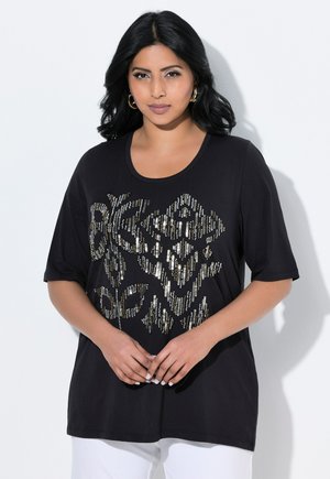Modelka nosí černé tričko s třpytivým grafickým designem, pózující na světlém pozadí, předvádějící módní ležérní look.