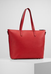 Červená kožená taška přes rameno se dvěma uchy, obdélníkový tvar, hladký povrch a zipem uzavíratelný vršek. Žádné viditelné vzory ani kovové detaily.