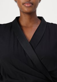 La blusa nera avvolgente presenta una trama liscia, un ampio colletto e una leggera lucentezza. Il design include una parte frontale incrociata per una silhouette aderente.