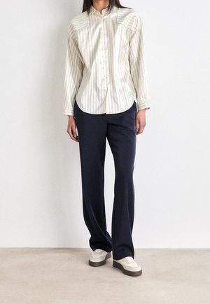 Femme portant une chemise claire à rayures boutonnée, un pantalon large bleu marine et des baskets blanches, debout contre un mur blanc uni.