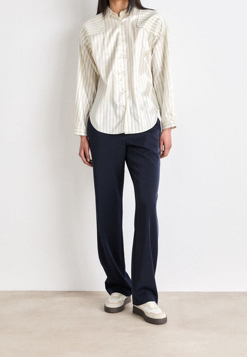 Femme portant une chemise claire à rayures boutonnée, un pantalon large bleu marine et des baskets blanches, debout contre un mur blanc uni.