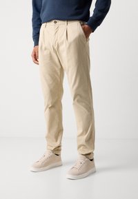 Beige cotton trousers with two front pleats, side pockets, and a single button closure, paired with light beige sneakers.