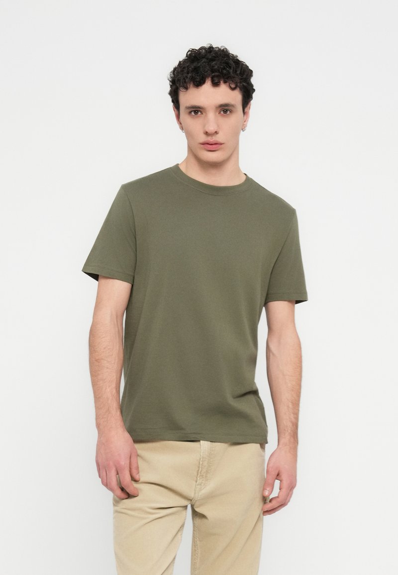 Βαμβακερό μπλουζάκι σε ελιά πράσινο με κοντά μανίκια, στρογγυλή λαιμόκοψη και εφαρμοστή γραμμή. Απαλή υφή, μινιμαλιστικό στυλ, χωρίς ορατά σχέδια.