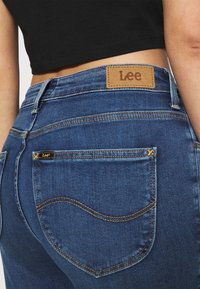 Mörkblå denimjeans med en brun läderetikett på bakre midjan, som har böjda sömnadsmönster och en liten gul accent.