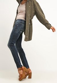 Olivově zelená oversized bunda, světle béžový top bez rukávů, tmavě modré skinny džíny a hnědé sandály na dřevěném podpatku s pásky kolem kotníků.