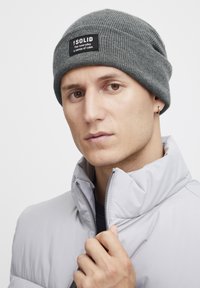 Gray knitted beanie with a black label featuring text, worn with a light gray zippered puffer jacket, showcasing a close-up view.