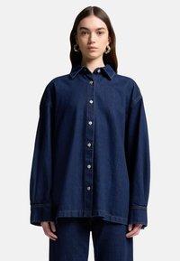 Mørkeblå denim oversized skjorte med krave, knapper foran og lange ærmer. Har synlige sømme og en afslappet pasform.