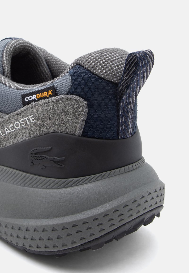 Männlicher grauer Sneaker aus unterschiedlichen Materialien, einschließlich strukturiertem Mesh und Filz-Akzenten, Gummisohle und markanter Branding seitlich.