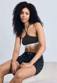 Μαύρο σουτιέν bralette με λεπτές τιράντες και λογότυπο Calvin Klein, σε συνδυασμό με μαύρο κοντό παντελονάκι με επαναλαμβανόμενο σχέδιο λογότυπου. Λείο υφασμάτινο υλικό.
