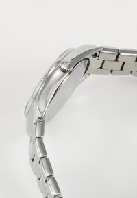 Klocka i silverfärgad rostfritt stål med polerad yta, rund klockkåpa och länkbandsrem. Har en texturerad krona och släta kanter.