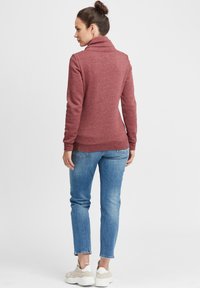 Un sweat-shirt bordeaux avec un col haut, coupe slim et poignets côtelés, associé à un jean bleu et des baskets blanches. Texture de tissu lisse.