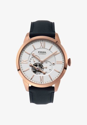 Montre automatique pour homme Fossil avec boîtier en or rose, cadran blanc, chiffres romains, engrenages visibles et bracelet en cuir noir.
