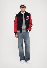 Jachetă varsity neagră și roșie cu detalii brodate, pulover gri și blugi lejere de culoare albastru deschis. Sportswear-uri albe completează ținuta.