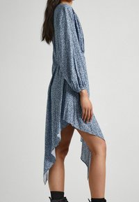 Sininen kukkamekko pitkillä hihoilla, epäsymmetrinen helma ja kevyttä kangasta, jossa on hienovarainen kuvio ja pehmeä rakenne.