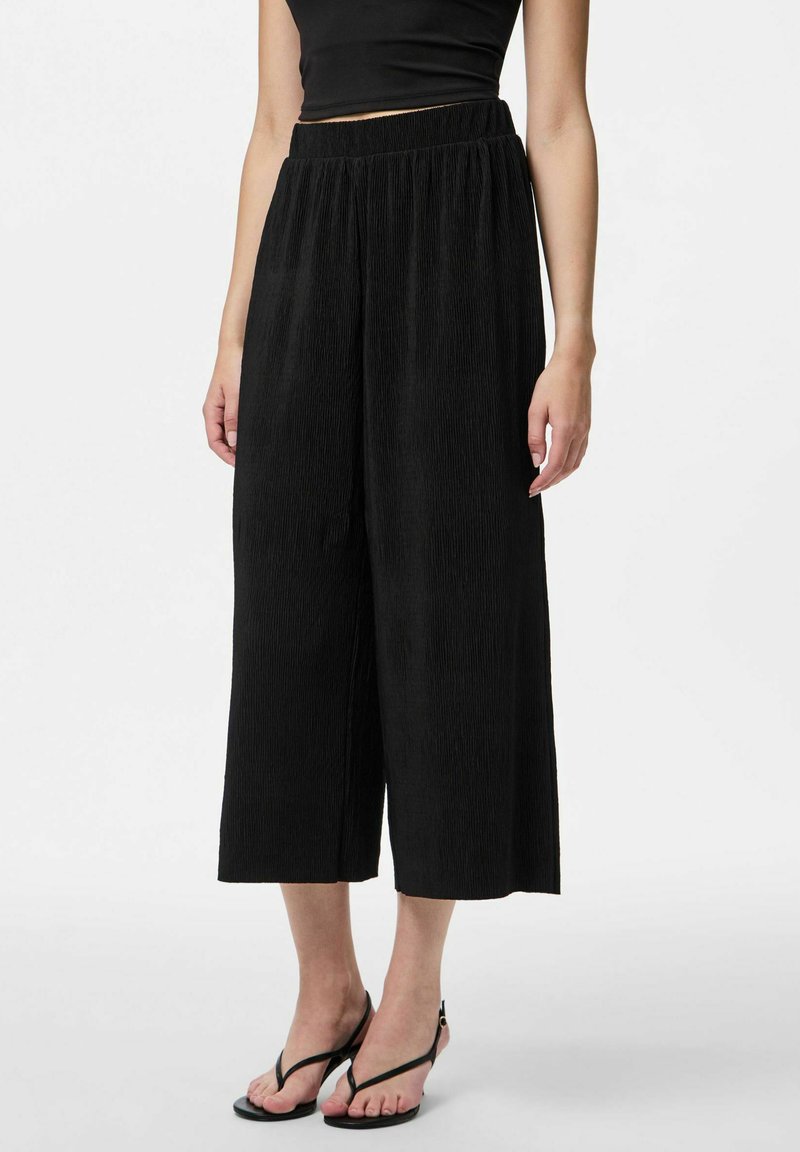 Svarta vida byxor med en texturerad, plisserad design. Elastisk midja för komfort. Matchas med tunna svarta sandaler på barfot.