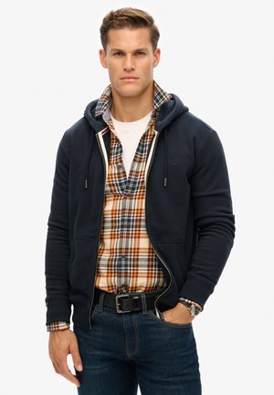 Navy zip-up hoodie over a plaid shirt with red, yellow, and blue tones. Features pockets, drawstring hood, and denim jeans.