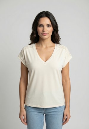 Jeune femme aux longs cheveux noirs portant un haut à manches courtes écru avec un col en V et un jean bleu clair, debout devant un fond gris uni.