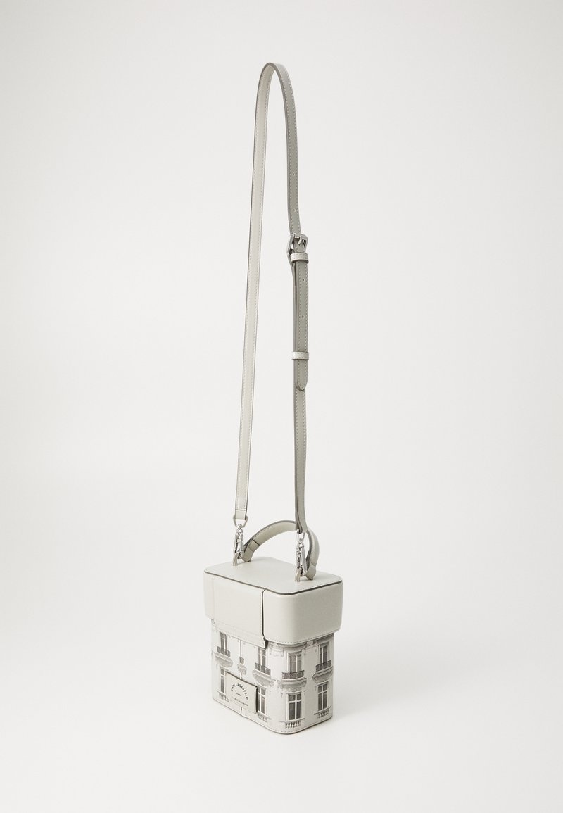 Sac bandoulière compact avec sangle ajustable, poignée supérieure et motif de façade de bâtiment sur la partie inférieure sur fond uni.