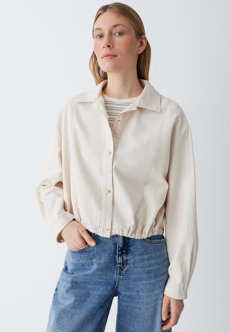 Vrouw draagt een lichtbeige jasje met knopen over een gestreept shirt, met één hand in de zak van een blauwe spijkerbroek, staand tegen een effen achtergrond.