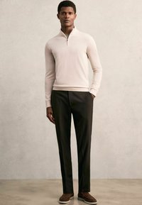 Helles beiges Zip-Up Sweatshirt, maßgeschneiderte schwarze Hose und braune Wildlederschuhe. Das Model posiert mit den Händen in den Taschen vor einem neutralen Hintergrund.