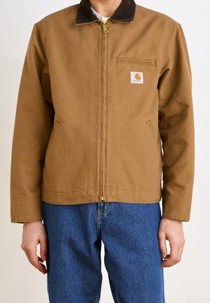 Mężczyzna w brązowej kurtce na zamek z czarnym kołnierzem i niebieskich dżinsach, stojący z ramionami swobodnie opuszczonymi wzdłuż ciała.