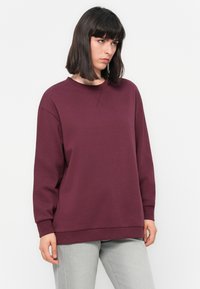 Burgundská mikina s kulatým výstřihem, dlouhými rukávy a žebrovanými manžetami. Vyrobena z měkké tkaniny s volným střihem, spárována se světle šedými kalhotami.
