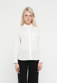 Bela srajca z dolgimi rokavi in ovratnikom, z gumbi, sprednjim žepom in sproščenim krojem. Iz lahkega blaga.