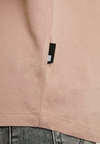 Chemise rose pâle à manches courtes avec une texture douce, ornée d'une étiquette noire et blanche à l'ourlet et de détails de couture visibles.