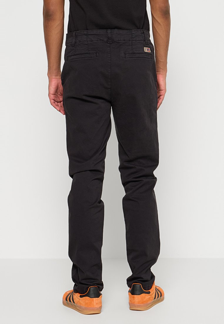 Schwarze Hosen mit schlanker Passform, ausgestattet mit Gesäßtaschen und einem kleinen Markenetikett; getragen mit orangefarbenen und schwarzen Turnschuhen.