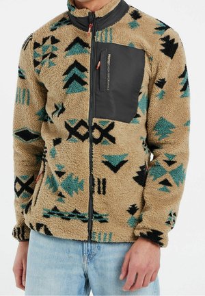 Mann trägt beige Fleecejacke mit schwarzen und türkisfarbenen geometrischen Mustern und einer schwarzen Reißverschlusstasche auf der Brust mit der Aufschrift "Protest High Performance."