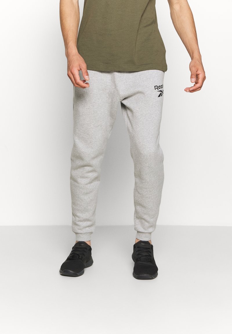 Pantalones deportivos grises con puños acanalados, con un logo negro de Reebok en el muslo izquierdo, combinados con zapatos deportivos negros y una camiseta verde oliva.
