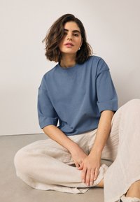 Modrá krátka mikina s okrúhlym výstrihom, v kombinácii so svetlo béžovými širokými ľanovými nohavicami. Modelka sediaca, ruky položené na kolenách.