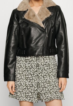Woman wearing black leather jacket with beige faux fur collar over a black and white floral dress, standing with arms relaxed by sides.